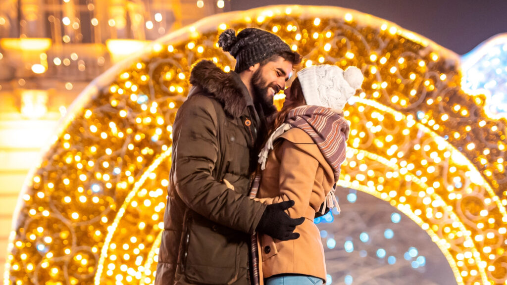 Liebeskolumne: Liebespaar umarmt sich vor leuchtendem Lichterbogen am Weihnachtsmarkt.
