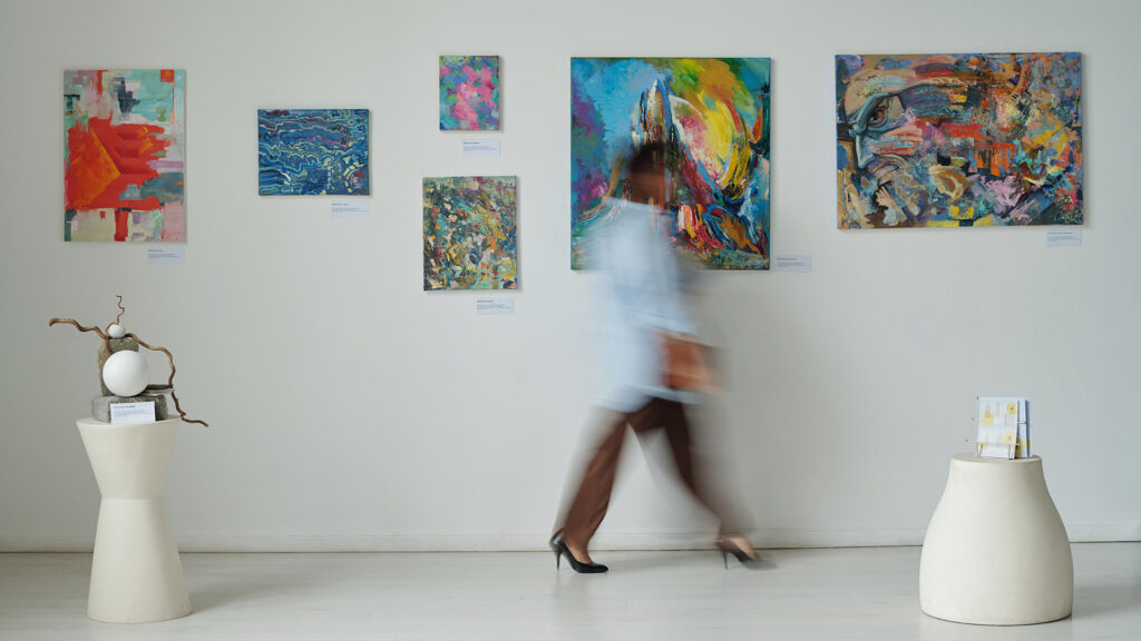 Kulturtipp: Verschwommene Bewegung einer jungen Frau, die an Gemälden an der Wand in einer Kunstgalerie vorbeigeht.