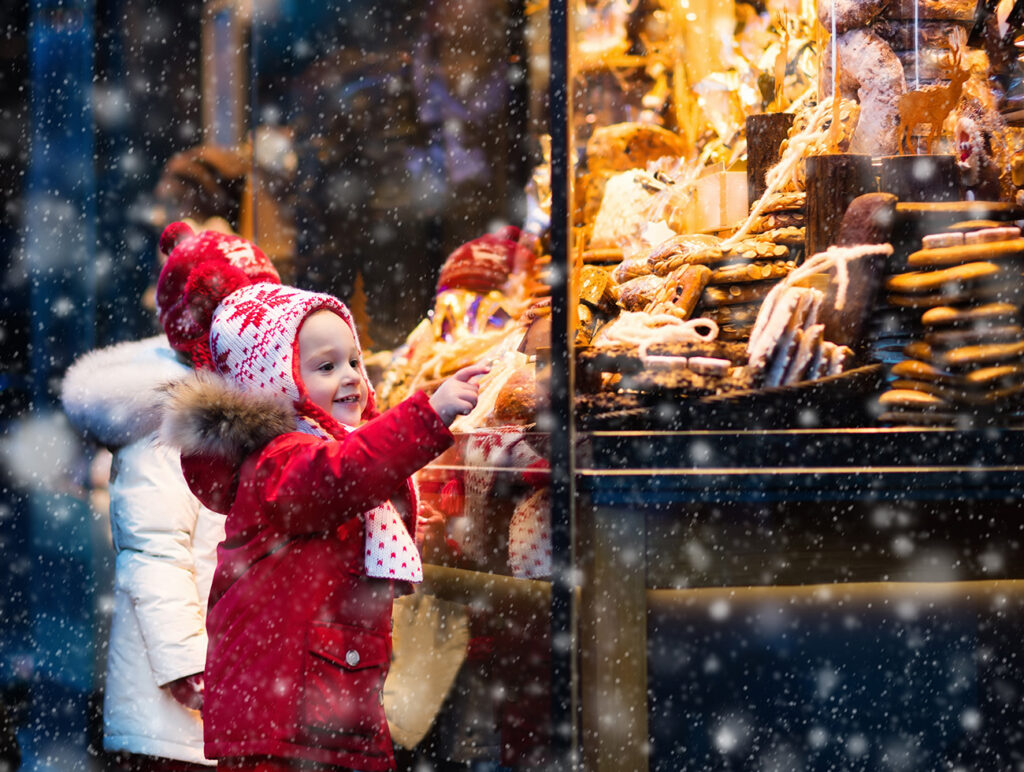 Adventmärkte: Kinder staunen über Süßigkeiten und Leckereien an einem weihnachtlichen Stand im Schneefall.