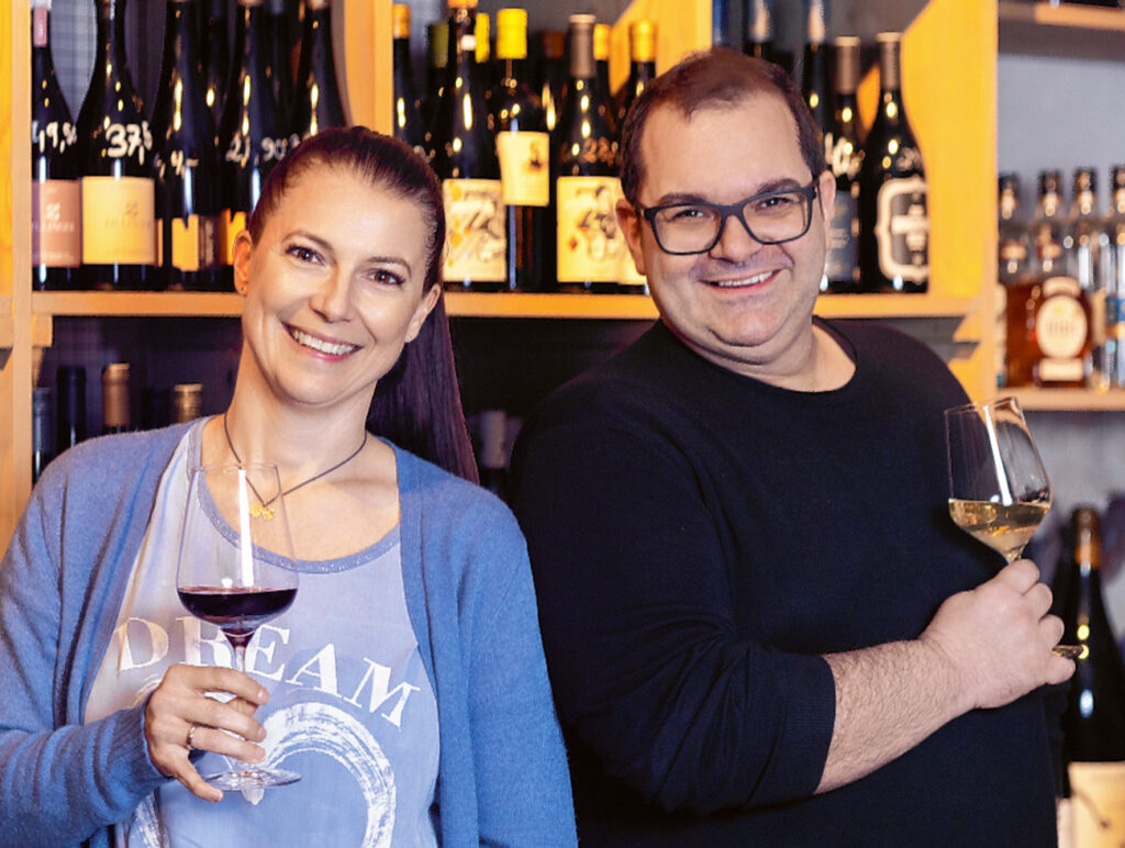 Zwei Personen stehen vor Weinregalen, beide halten Weingläser und lächeln in die Kamera.