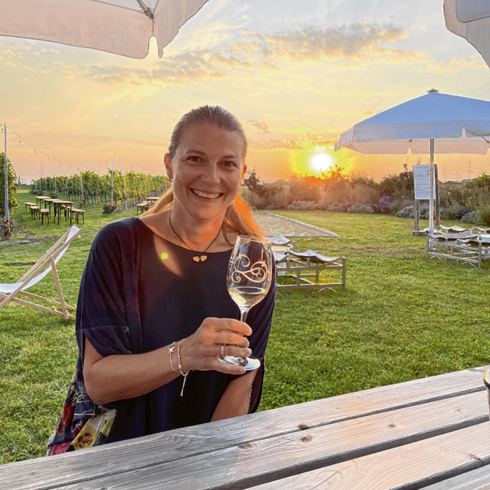 Corni Winelover: Frau sitzt an einem Holztisch mit Weinglas in der Hand, im Hintergrund Weinberge und Sonnenuntergang.