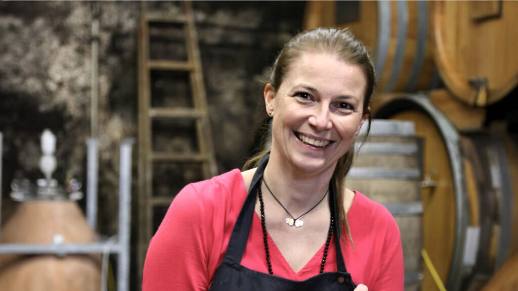 Corni Winelover: Frau mit rosa Oberteil und Schürze lächelt in einer Weinkellerei, im Hintergrund Holzfässer und Leiter.