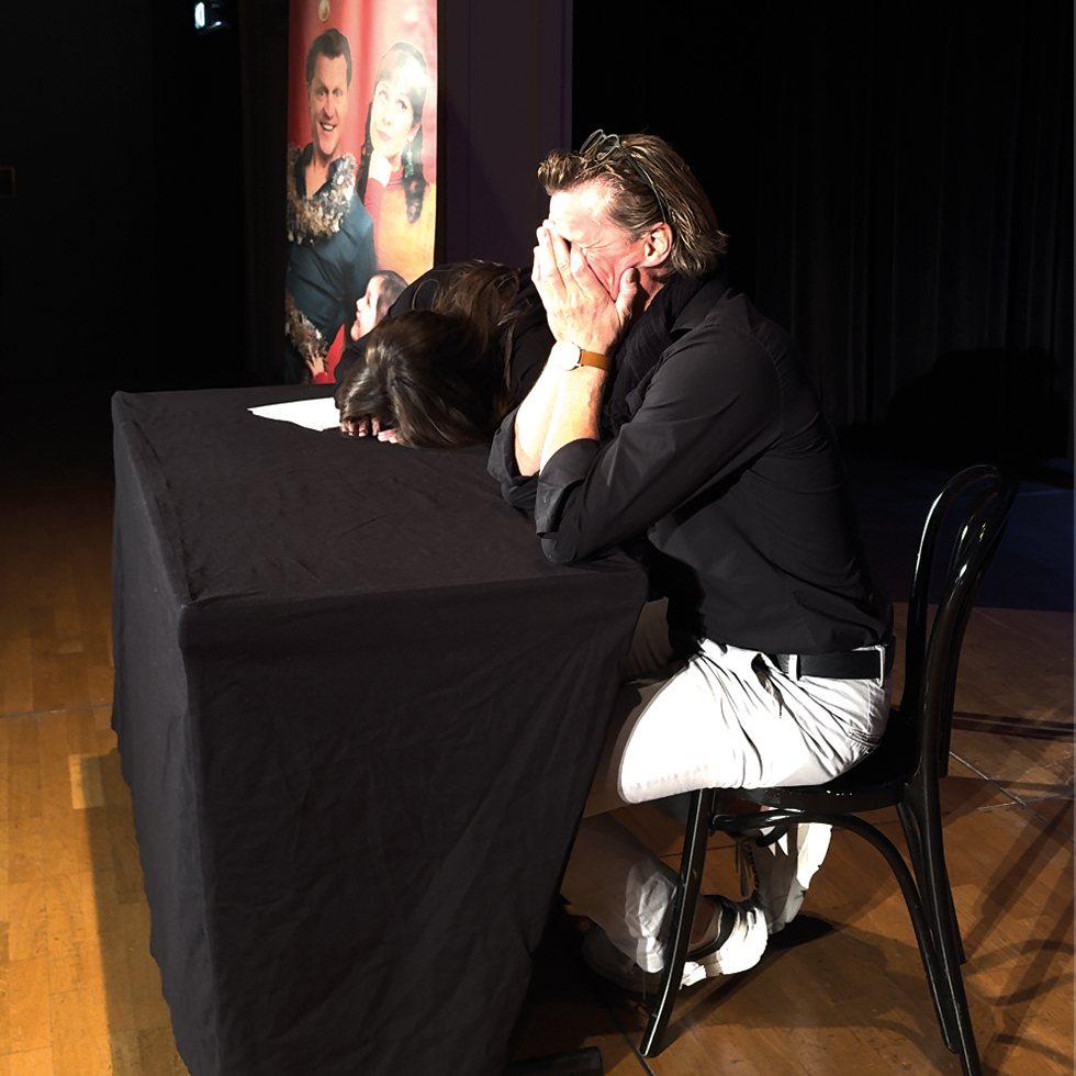 Zwei Personen lehnen sich lachend über einen Tisch auf der Bühne, im Hintergrund ein Poster.