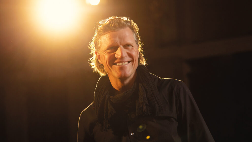 Gery Seidl: Mann lächelt in warmem Bühnenlicht, trägt dunkles Hemd und Schal.