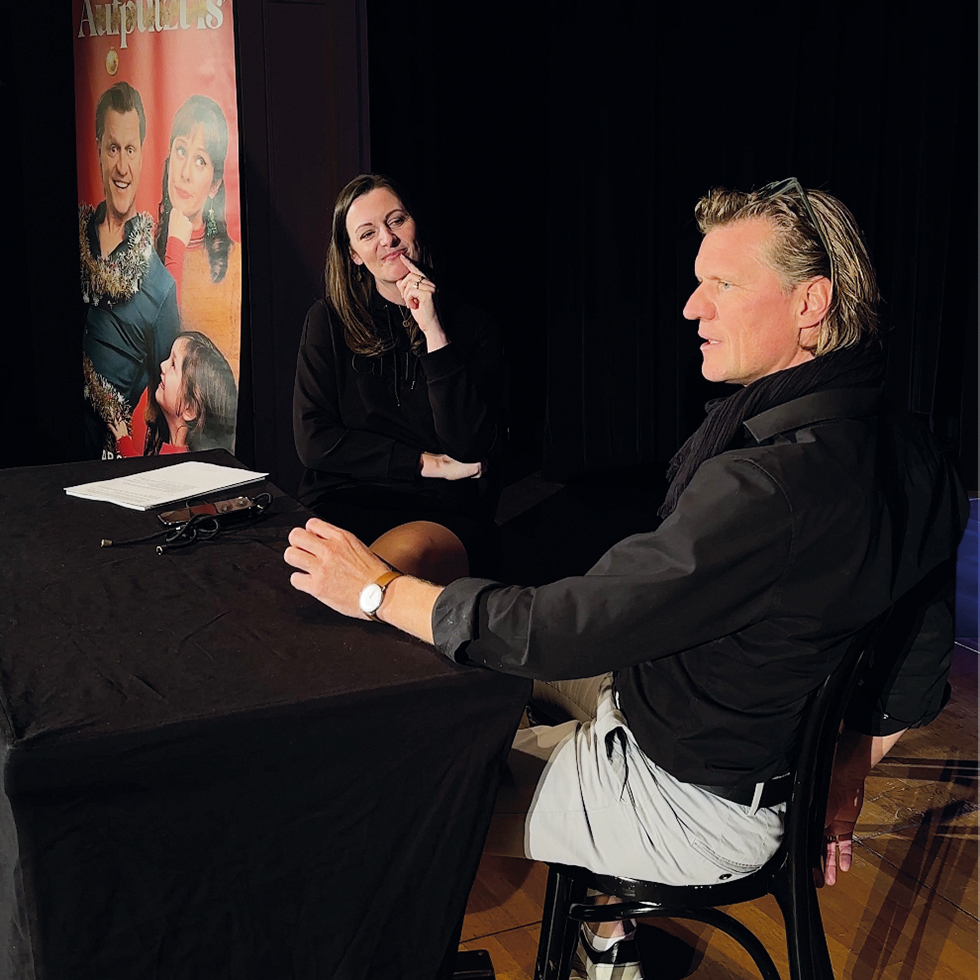 Gery Seidl: Zwei Personen sitzen an einem Tisch auf einer Bühne und unterhalten sich, im Hintergrund ein Poster.