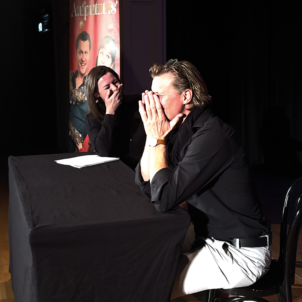 Zwei Personen an einem Tisch auf einer Bühne lachen und halten sich vor Lachen das Gesicht.