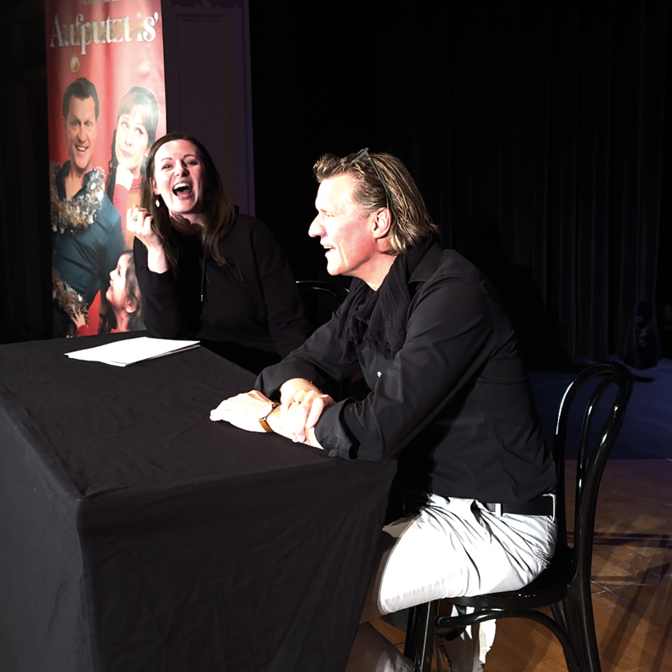 Zwei Personen sitzen auf einer Bühne und lachen während eines Gesprächs.