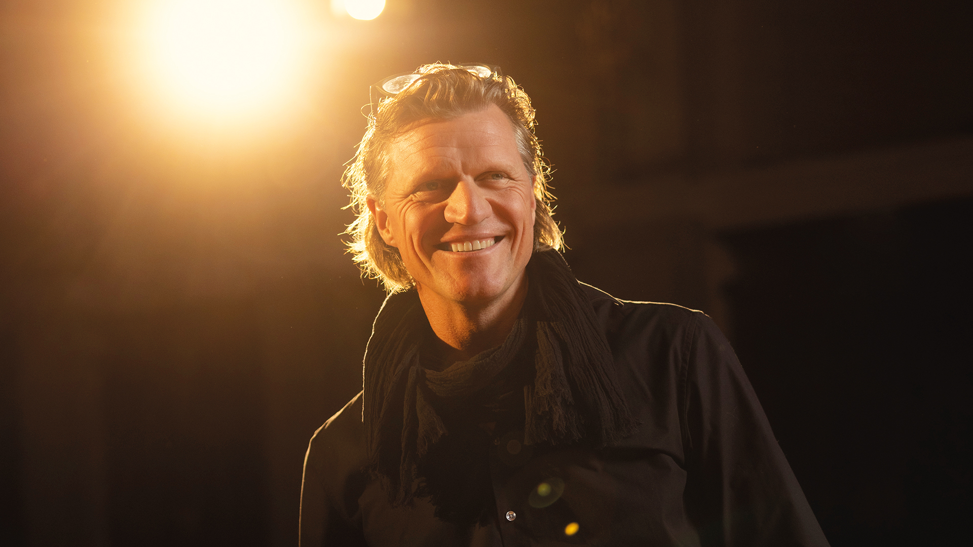 Gery Seidl: Mann lächelt in warmem Bühnenlicht, trägt dunkles Hemd und Schal.