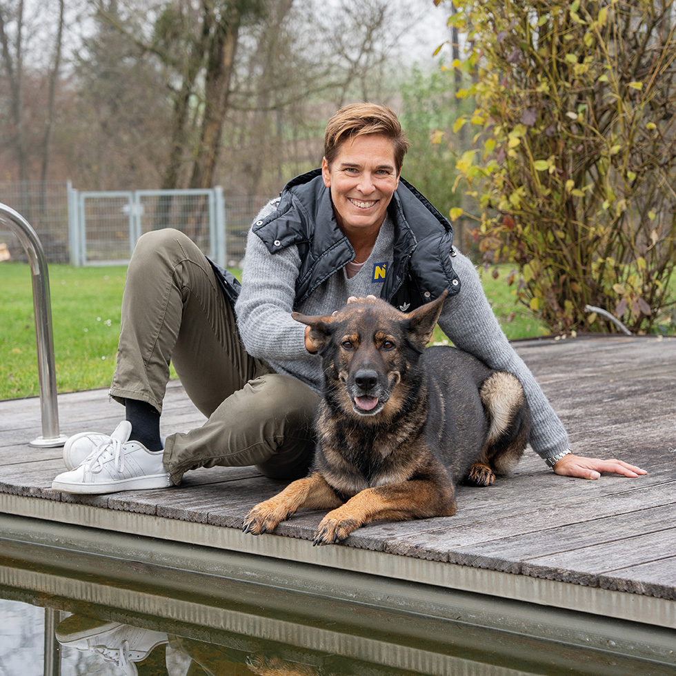 Michaela Dorfmeister: Frau sitzt am Holzsteg neben einem Hund an einem Gartenteich.