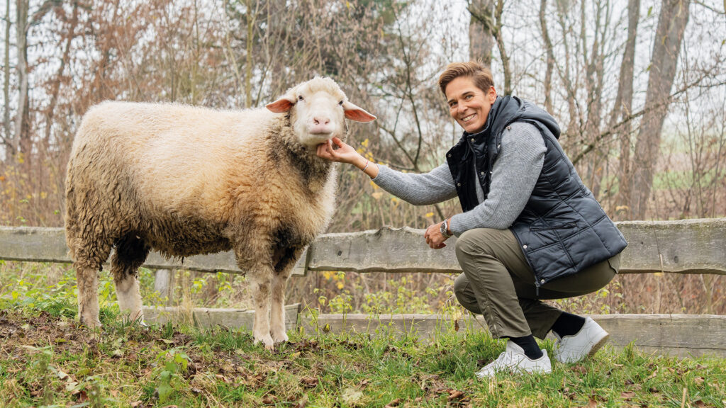 Michaela Dorfmeister: Frau kniet neben einem Schaf auf einer Wiese.
