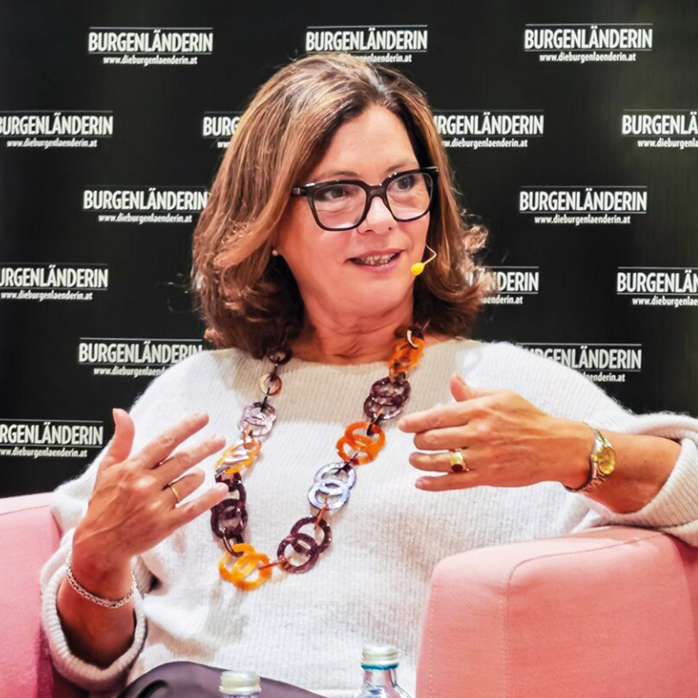 Podiumsdiskussion: Rednerin mit Headset auf einem rosa Sessel vor Veranstaltungs-Backdrop