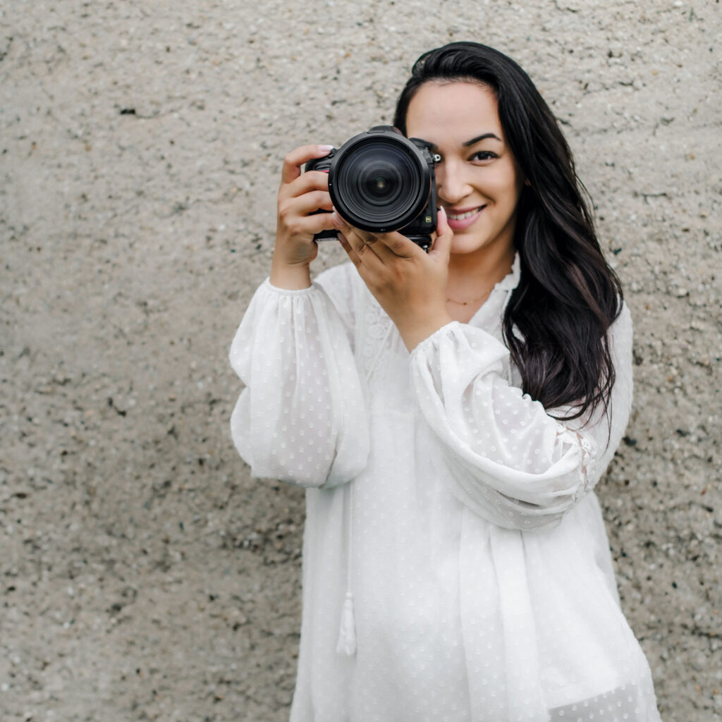 Eine Fotografin im weißen Shirt schaut durch ihre Kamera und lächelt dabei.