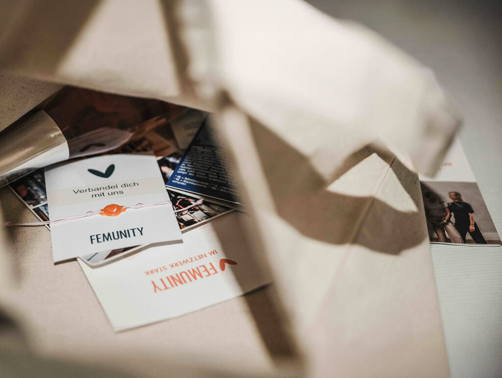 Nahaufnahme eines FEMUNITY-Armbands und Infomaterials in einer Stofftasche.