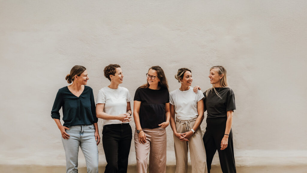 Femunity: Fünf Frauen stehen lächelnd nebeneinander vor einer hellen Wand.“