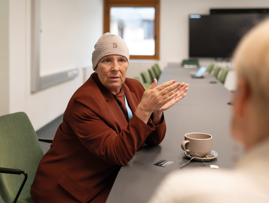 Person im braunen Blazer gestikuliert während eines Gesprächs in einem Konferenzraum.