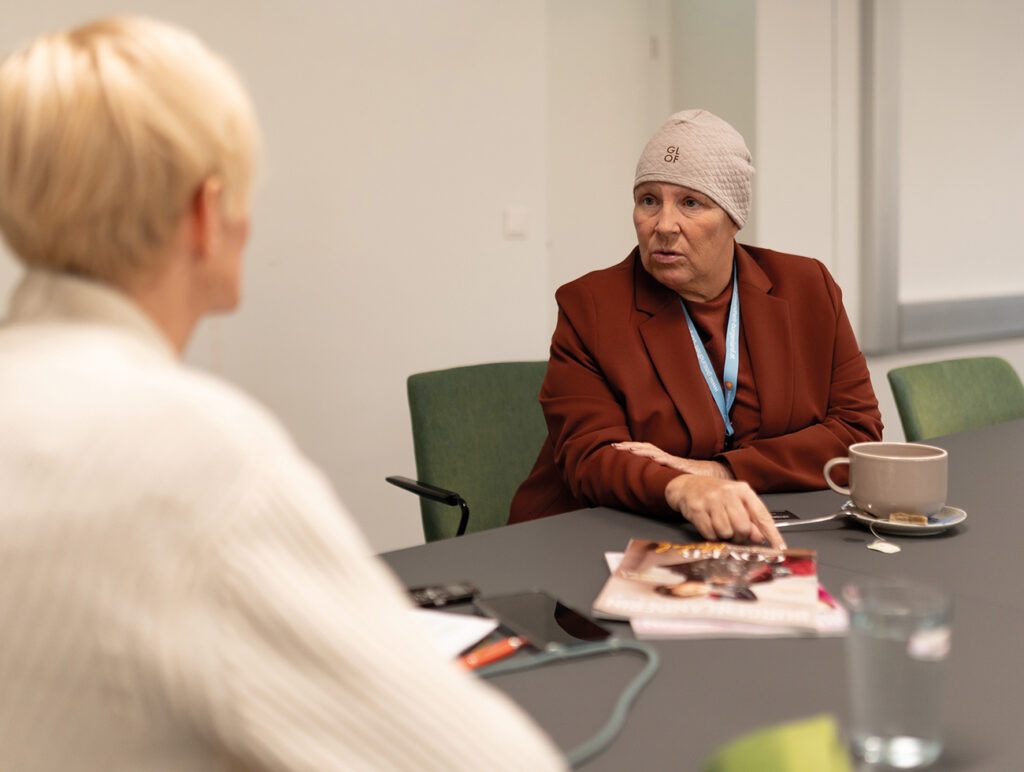 Person im braunen Blazer spricht während eines Interviews und zeigt auf Magazine am Tisch.