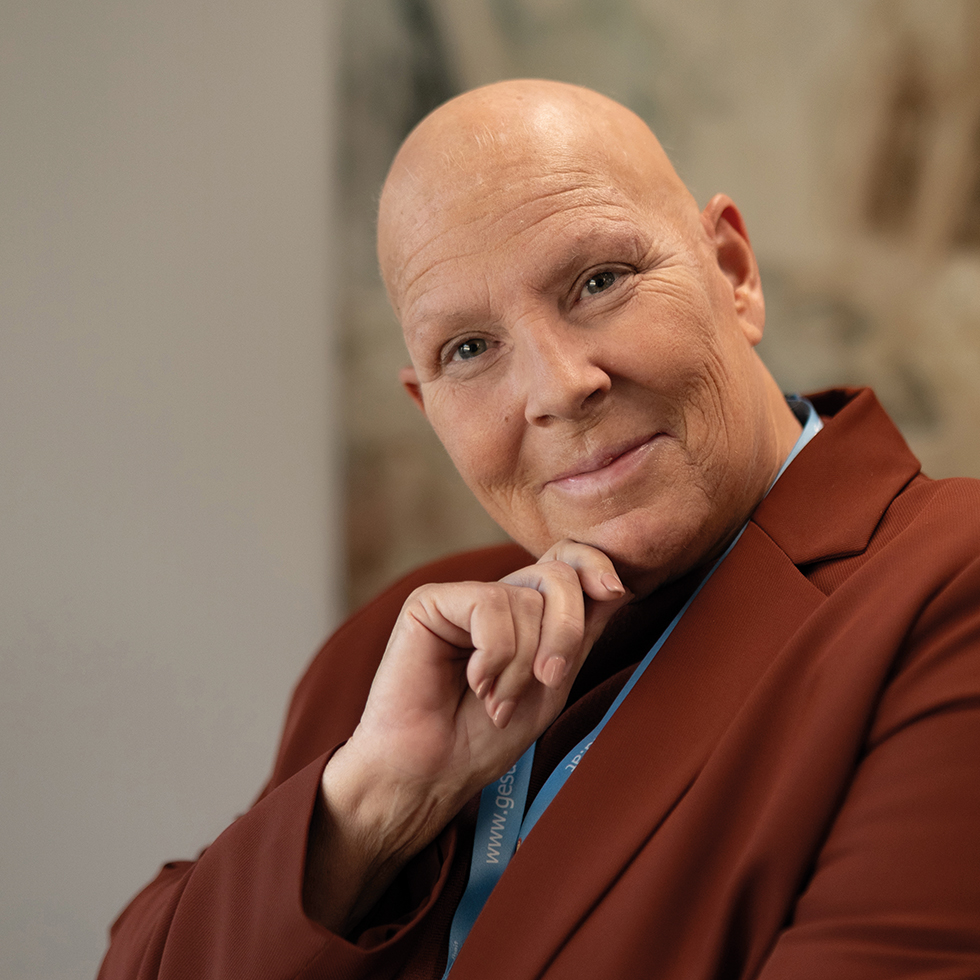 Porträt einer lächelnden Frau mit Glatze in braunem Blazer, die den Kopf auf die Hand stützt.