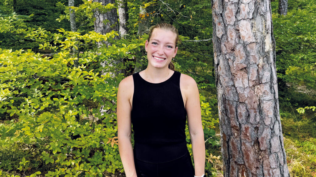 Absolventin: Eine junge Frau steht lächelnd in einem sonnigen Wald, umgeben von frischem Grün und Bäumen.