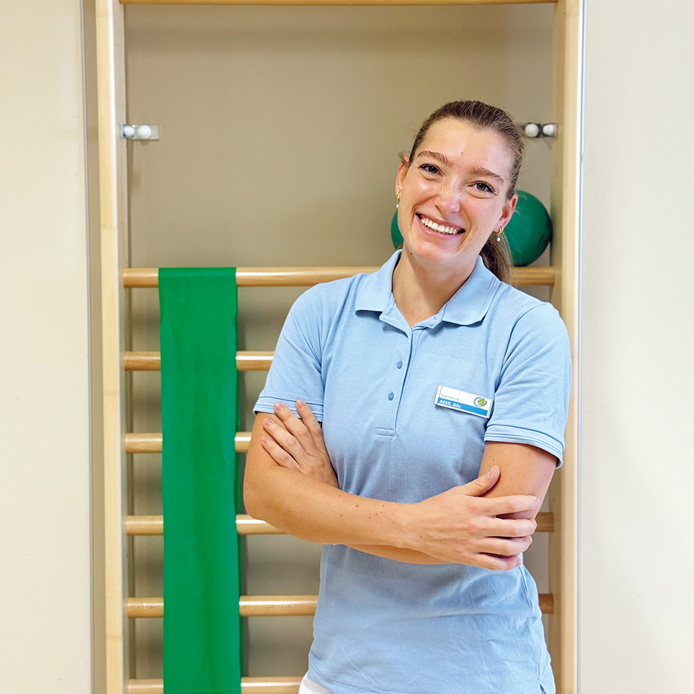 Absolventin: Eine Physiotherapeutin steht lächelnd vor einer Sprossenwand und wirkt freundlich und zugewandt.