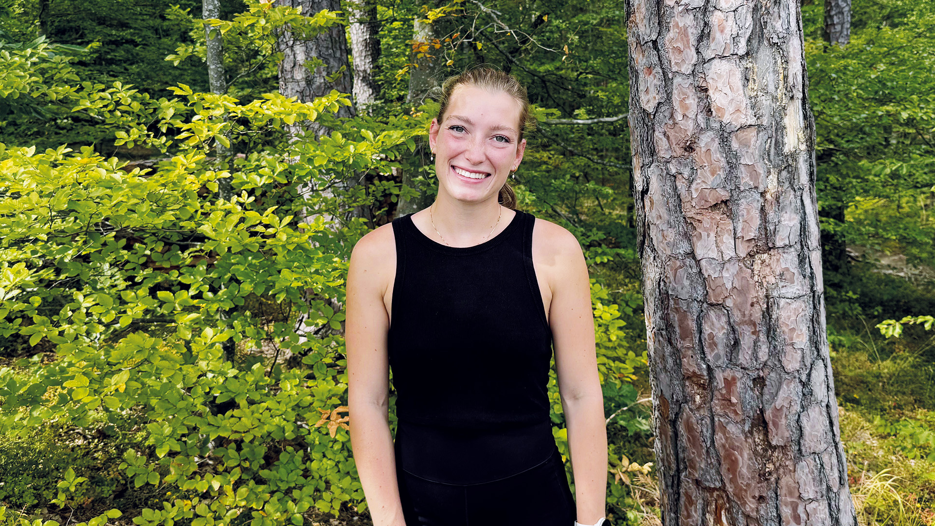 Absolventin: Eine junge Frau steht lächelnd in einem sonnigen Wald, umgeben von frischem Grün und Bäumen.