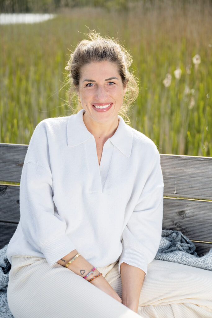 Selbstfürsorge: Porträt einer lächelnden Frau auf einer Holzbank vor hohem Schilf im warmen Sonnenlicht.