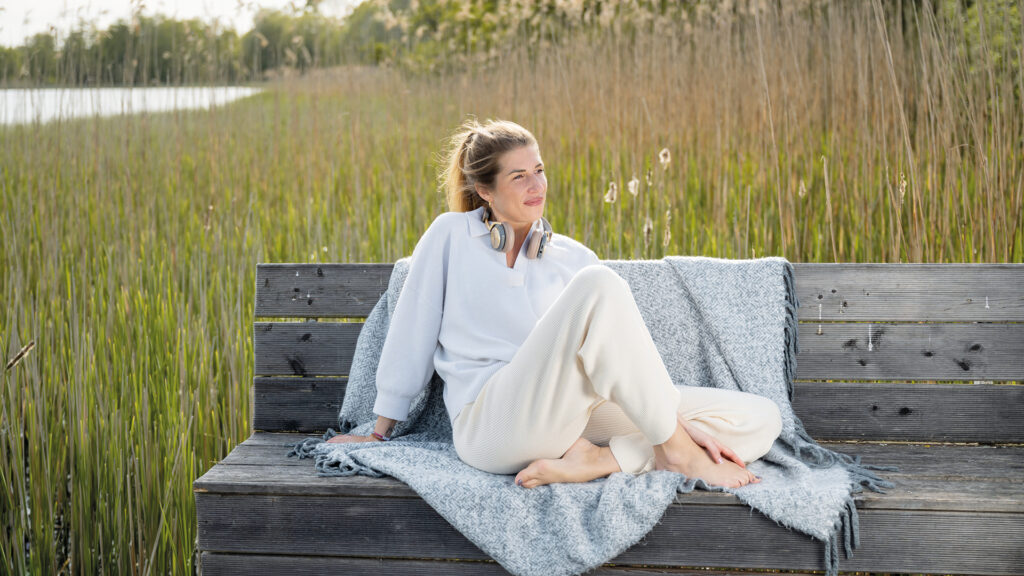 Selbstfürsorge: Frau sitzt entspannt auf einer Holzbank am See, eingehüllt in eine Decke und mit Kopfhörern um den Hals.
