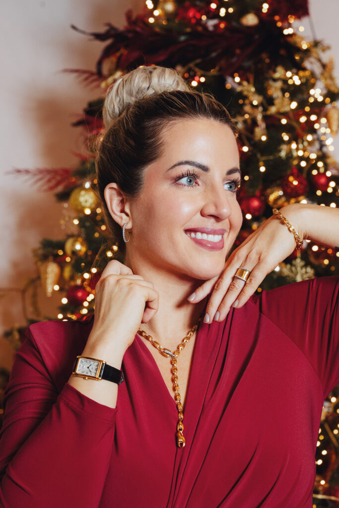 Frau in elegantem roten Kleid präsentiert goldenen Schmuck vor einem festlich geschmückten Weihnachtsbaum.