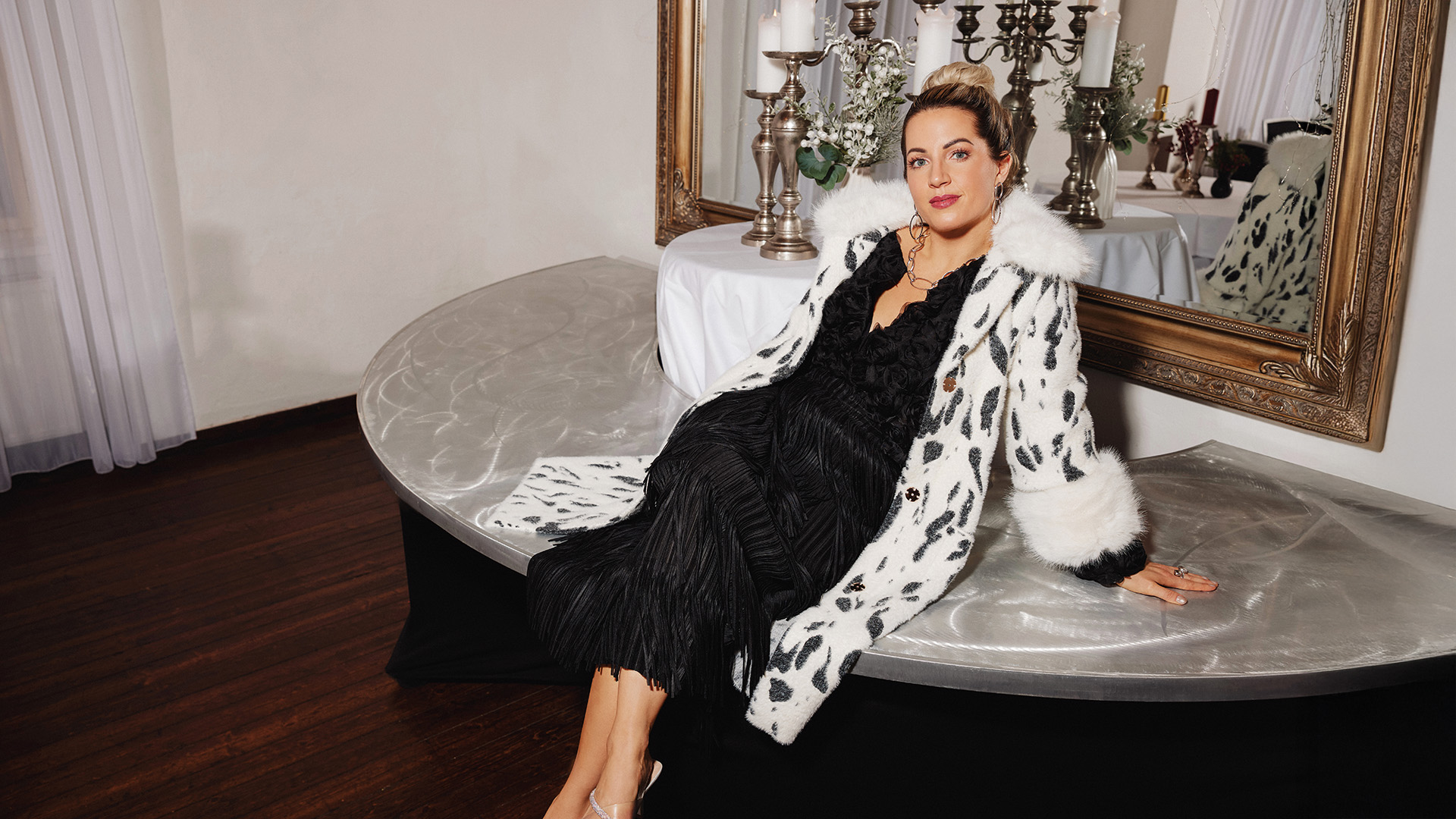 Abendlooks: Frau in schwarz-weißer Felljacke posiert elegant auf einer silbernen Bank vor einem großen Spiegel.