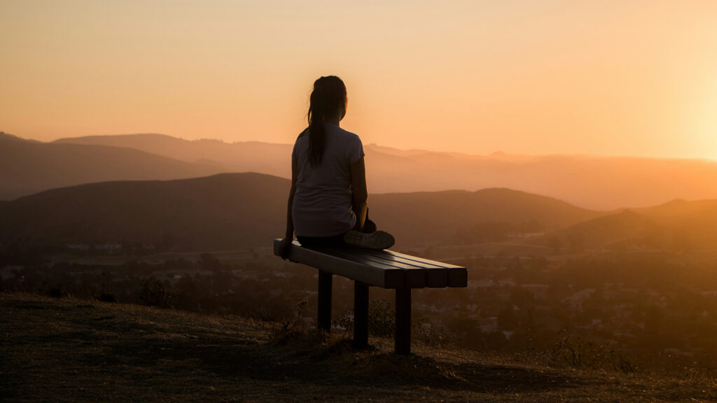 Person sitzt auf einer Bank und blickt bei Sonnenuntergang über eine hügelige Landschaft, die in warmes Abendlicht getaucht ist.“