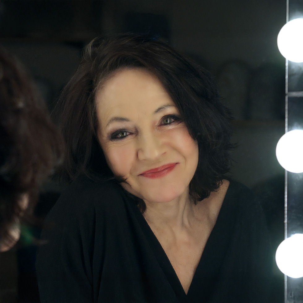 Maria Bill: Porträt einer dunkelhaarigen Frau mit freundlichem Lächeln vor einem beleuchteten Spiegel.