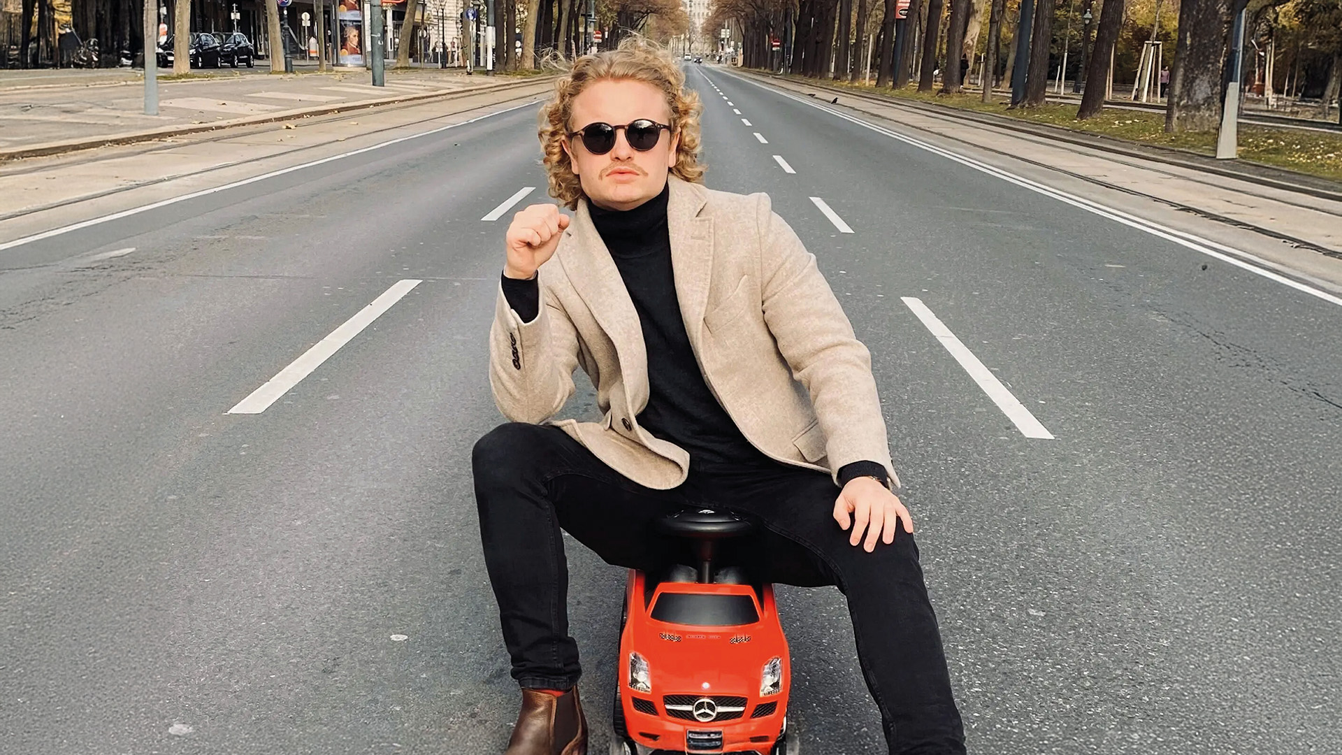 Februar Freizeittipps: Mann mit Sonnenbrille posiert auf einem roten Spielauto auf einer breiten Straße.