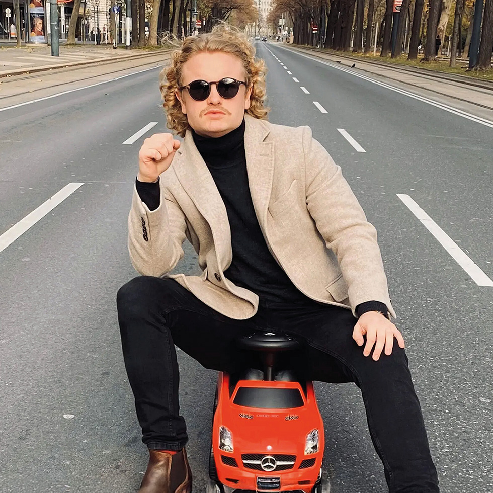 Februar Freizeittipps: Mann sitzt auf einem roten Spielauto mitten auf einer leeren Straße.