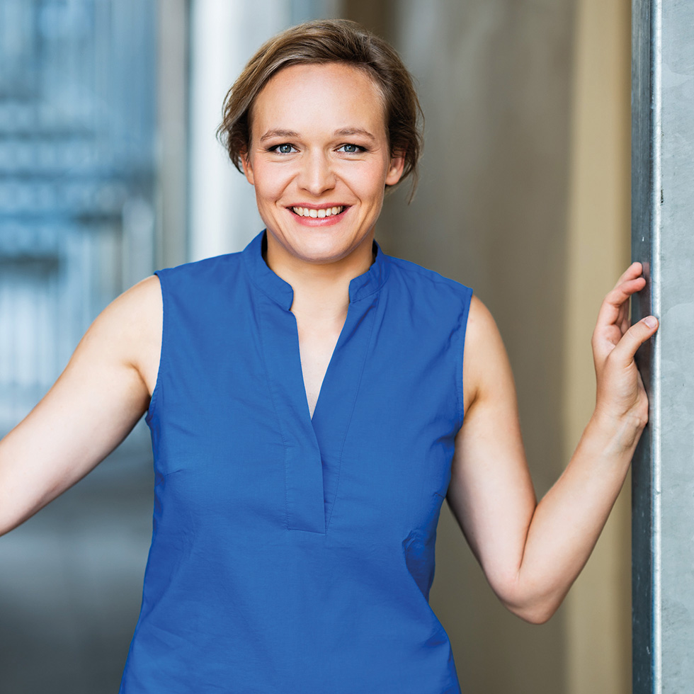 Freizeittipp: Frau im blauen ärmellosen Kleid steht lächelnd in einem hellen Durchgang.