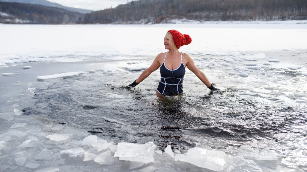 Wellnesstrends: Person steht bis zur Taille im kalten See, trägt Badebekleidung und eine rote Mütze, umgeben von Eis am Ufer.