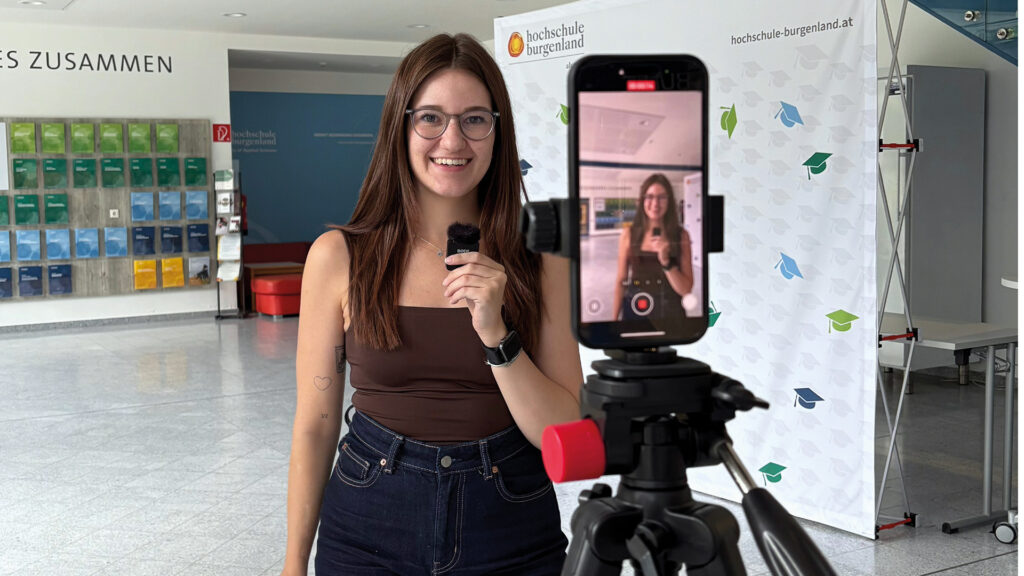 Lena Sagmeister: Junge Frau mit langen rot-braunen Haaren und brauner Brille filmt sich selbst mit einen Mobiltelefon auf einem Stativ.