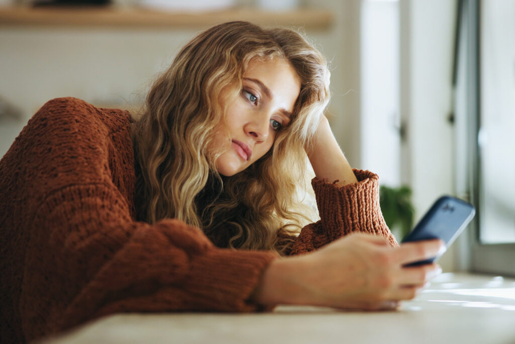 Blonde Frau lehnt gelangweilt mit einer Hand am Kopf und Handy in der 2. Hand auf einer Tischplatte