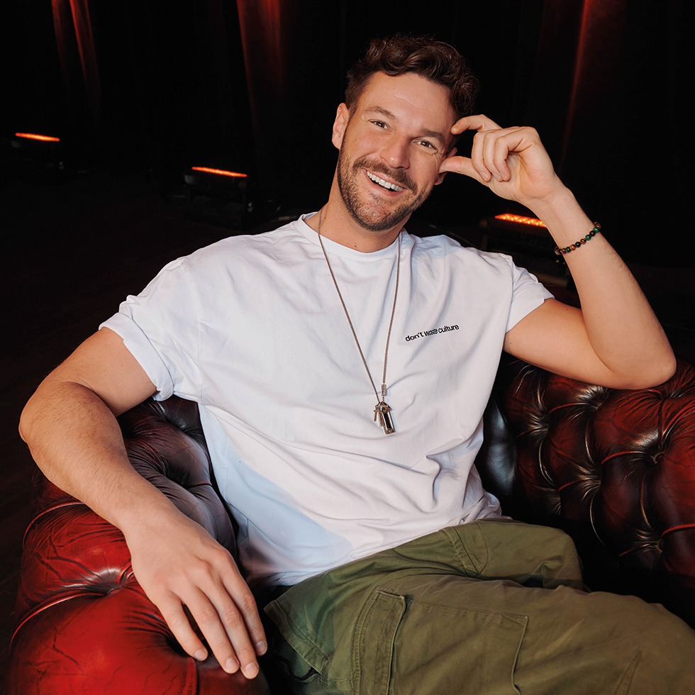 Hawi D'Ehre: Paul Pizzera sitzt auf einem Couchsessel und blickt lachend, auf einer Hand abgestützt in die Kamera