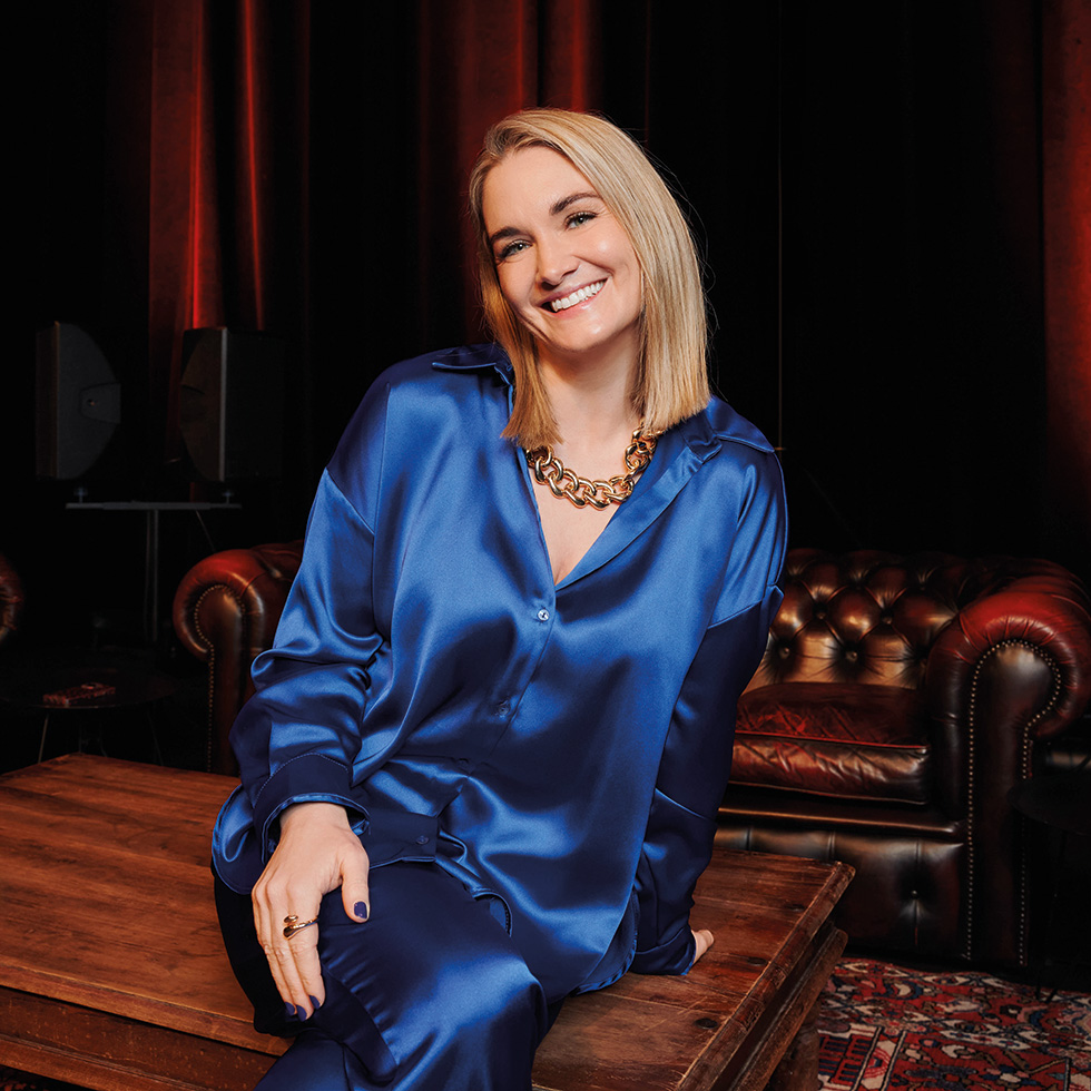 Hawi D'Ehre: Gabi Hiller – eine blonde Frau im blitzblauem Satin-Anzug lächeld sitzend in die Kamera