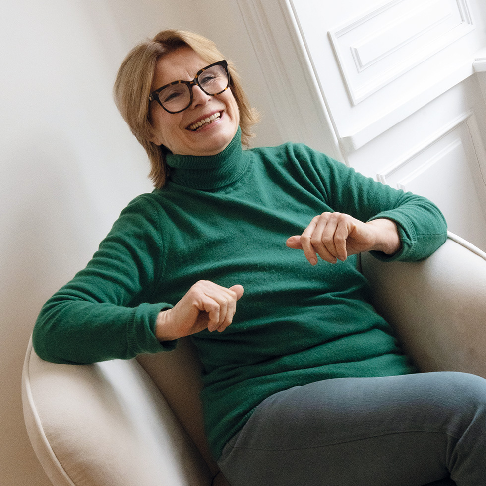 Edit Schlaffer: Frau mit Brille und grünem Rollkragenpullover sitzt lachend auf einem Sessel