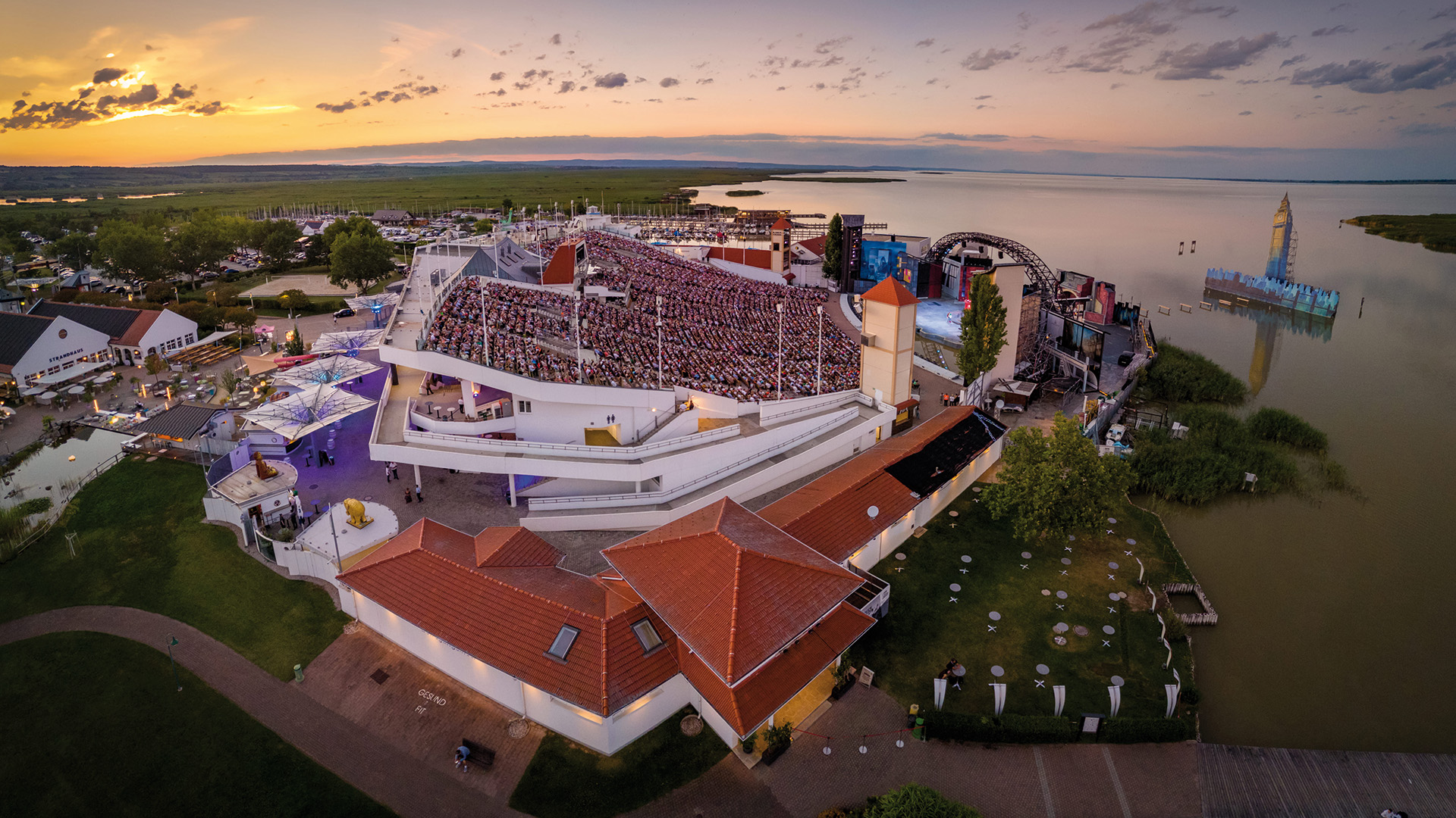 #bgld: Luftaufnahme einer großen Seebühne mit vollbesetztem Zuschauerraum bei Sonnenuntergang