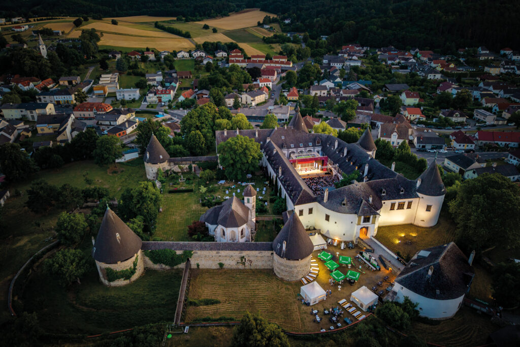 #bgld: Luftaufnahme einer Burganlage mit Festivalbetrieb in der Abenddämmerung