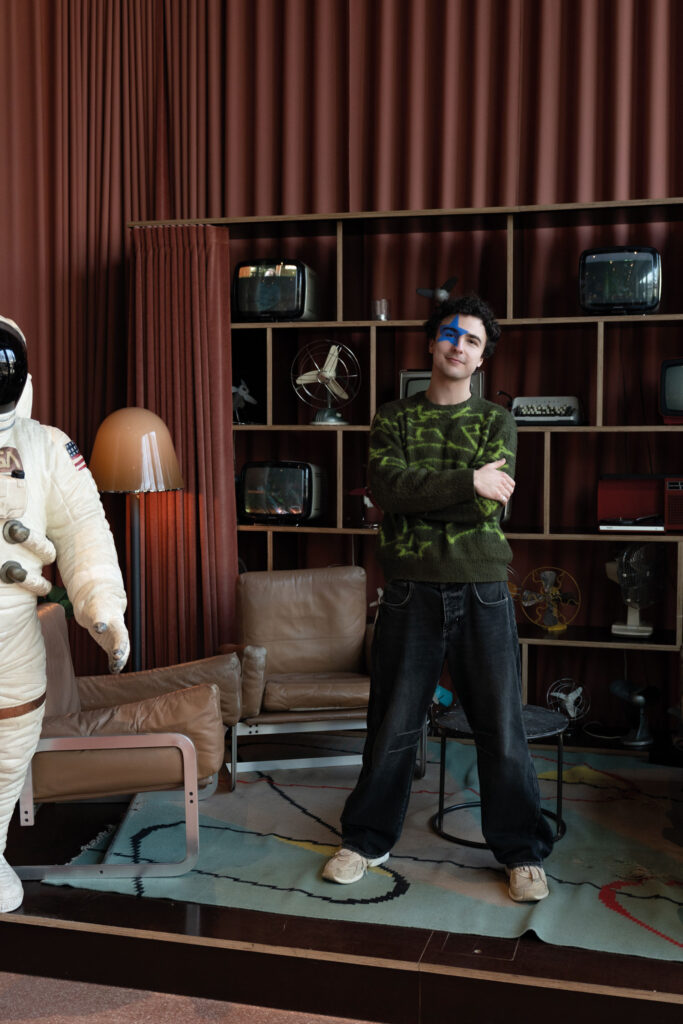 Portrait von COSMÓ in einem Wohnraum mit alten Monitorem und einem Astronautenanzug.