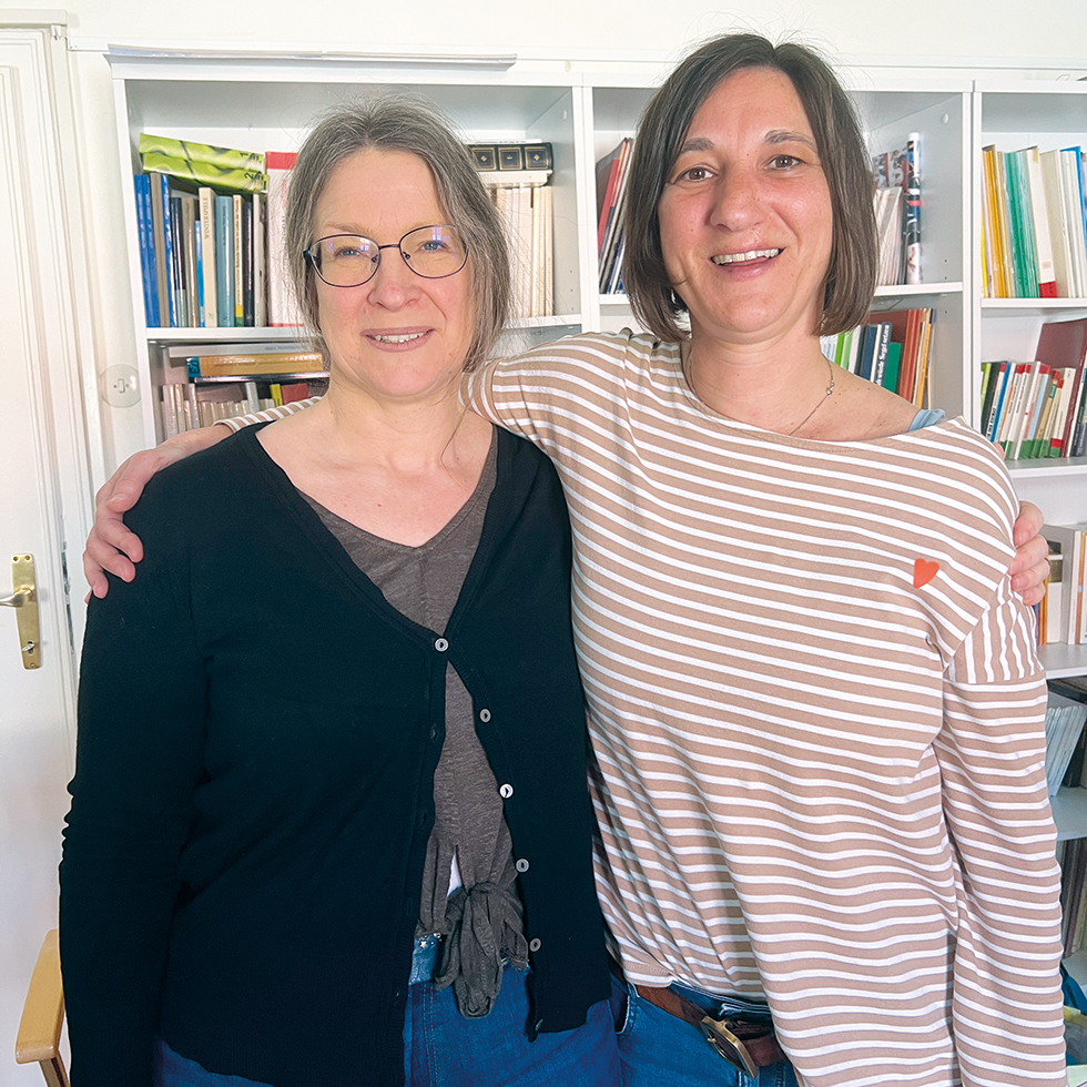 Letzte Hilfe: Zwei Frauen stehen umarmt vor Bücherregal und lächeln in die Kamera