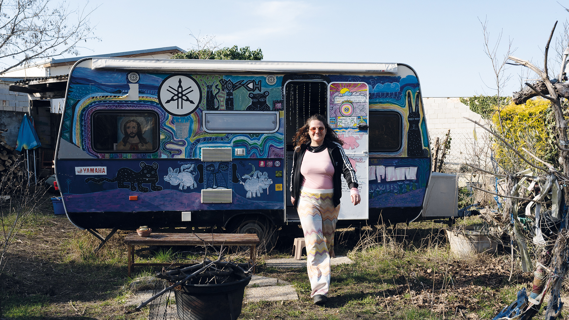 Mut: Frau geht aus einem bunt bemalten Wohnwagen in einem Garten heraus