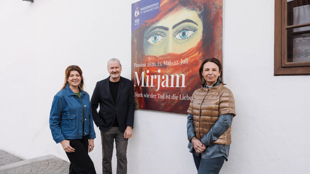 Passion: Drei Personen stehen vor einem Plakat der Passionsspiele „Mirjam“