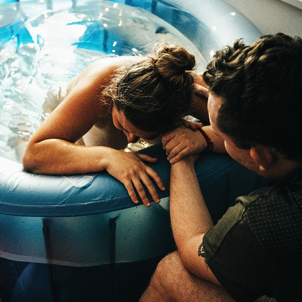 Eine Frau stützt sich während der Geburt auf einen aufblasbaren Pool, während eine Begleitperson ihre Hand hält.
