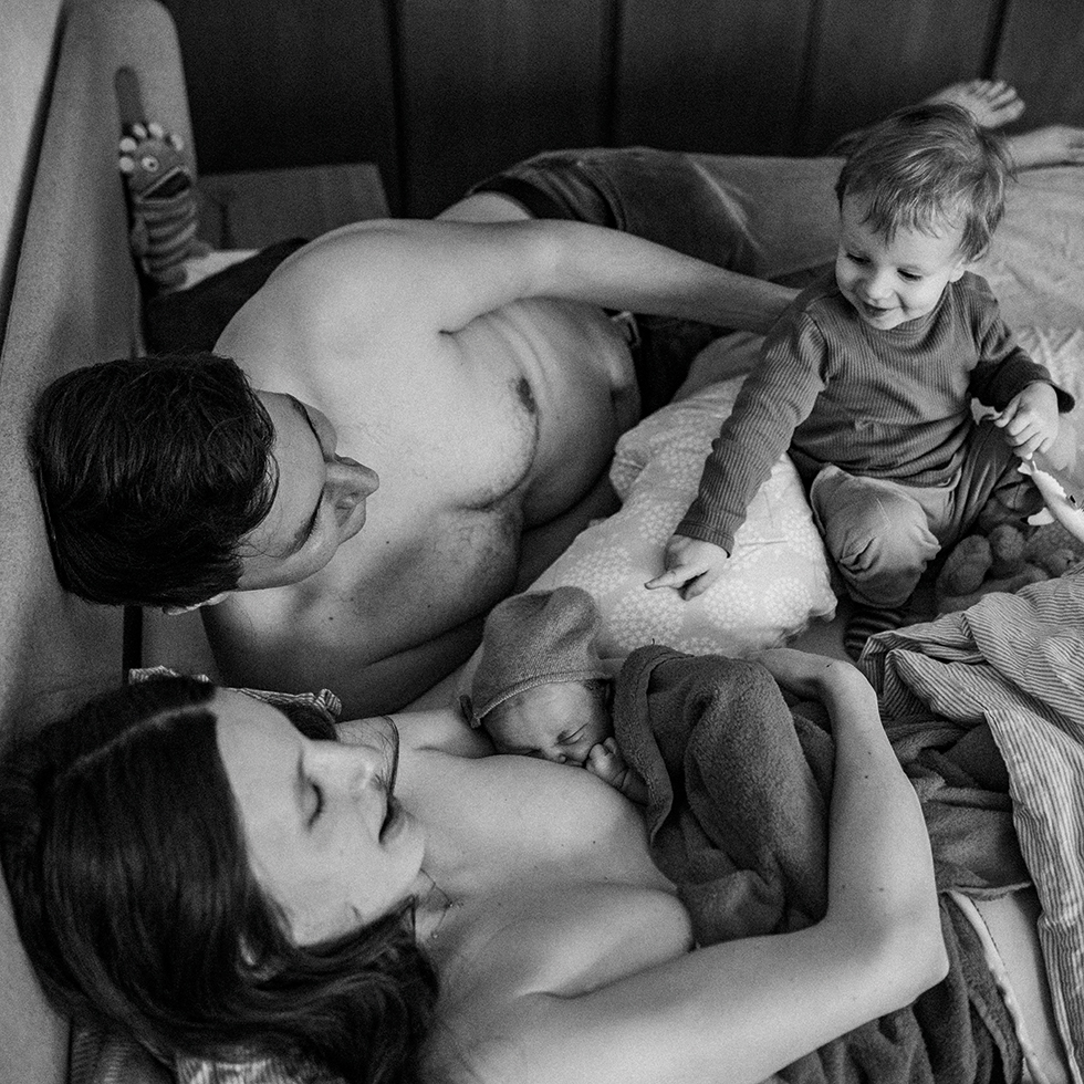 Eine Familie liegt gemeinsam im Bett, während ein Neugeborenes gestillt wird und ein Kleinkind daneben sitzt.