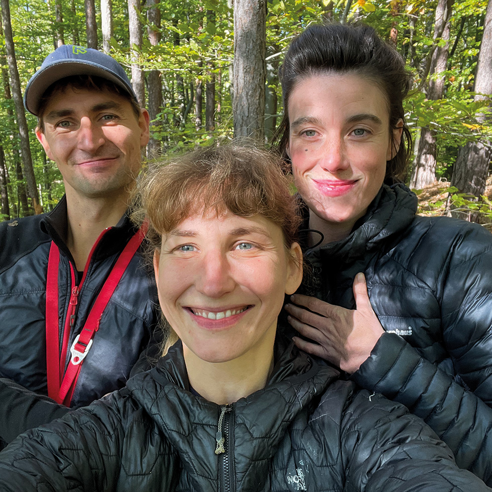woman/mOther: Drei Erwachsene machen ein Selfie im Wald, tragen Outdoor-Kleidung und lächeln in die Kamera.
