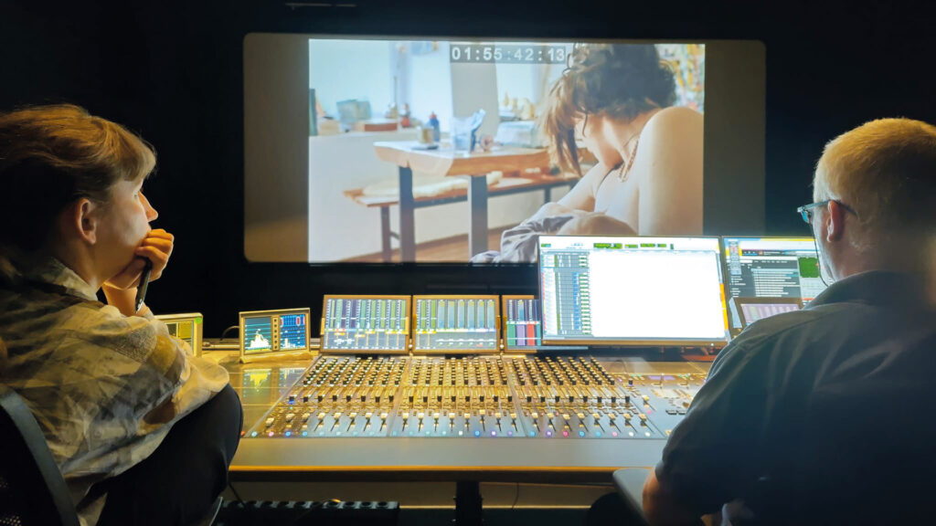 woman/mOther: Zwei Personen sitzen im Tonstudio vor Mischpulten und betrachten eine Filmszene einer stillenden Mutter auf einer großen Leinwand.