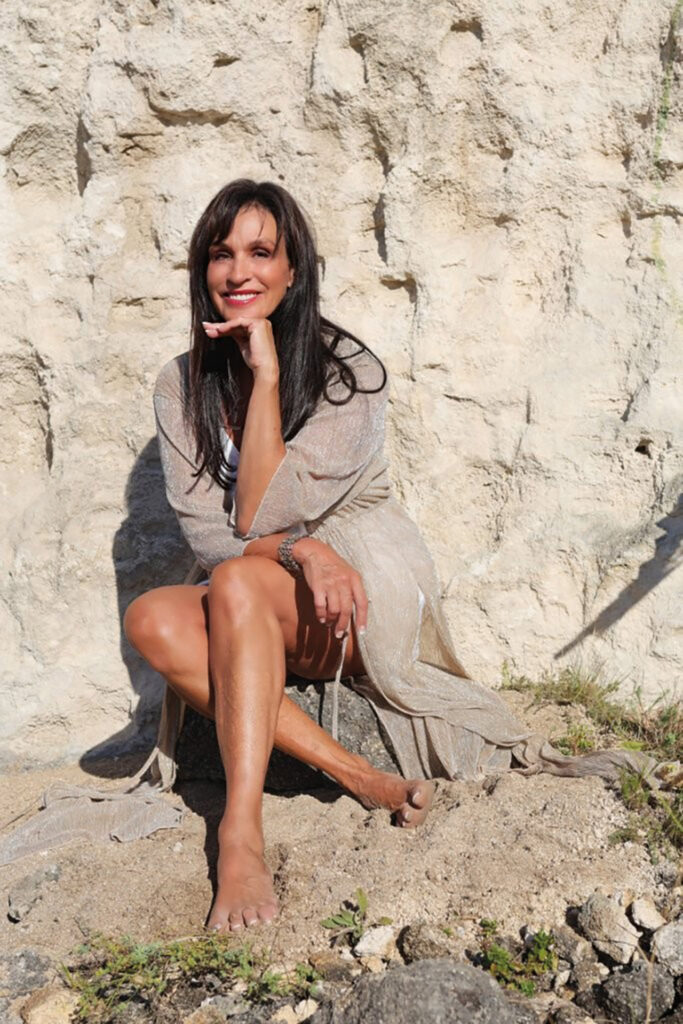 Catherine Sica sitzt barfuß im Sonnenlicht vor einer hellen Felswand und posiert entspannt mit einem leichten, beigen Kleid.